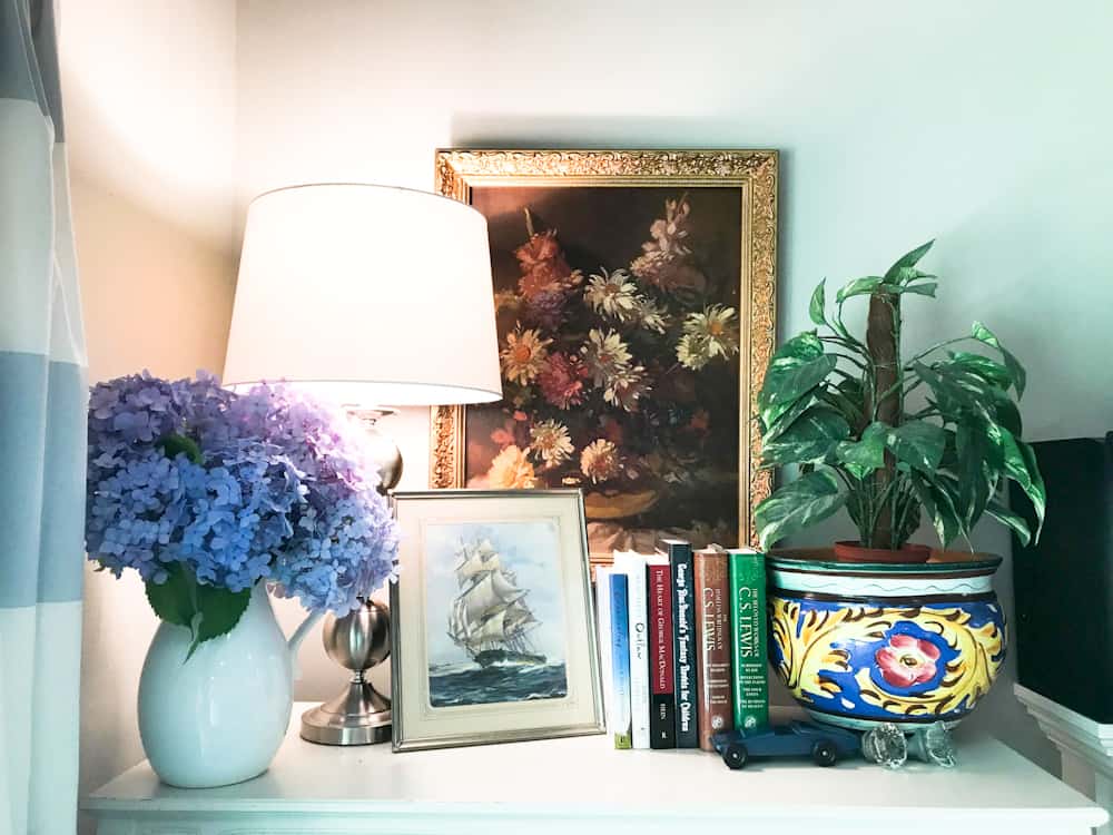 Life Full and Frugal / Summer Living Room Makeover / Top of Book shelf decorated with books, a vintage print of a ship on open water, a floral painting, a white vase with blue hydrangeas and a plant nestled in Mexican pottery