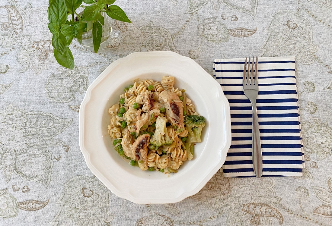 cajun chicken pasta with brocolli and green peas served in a white bowl with a blue and white striped cloth napkin placed beside it, a fork placed on the napkin, patterned table cloth and a small glass of fresh basil - Life Full and Frugal - Easy Cajun Chicken Pasta