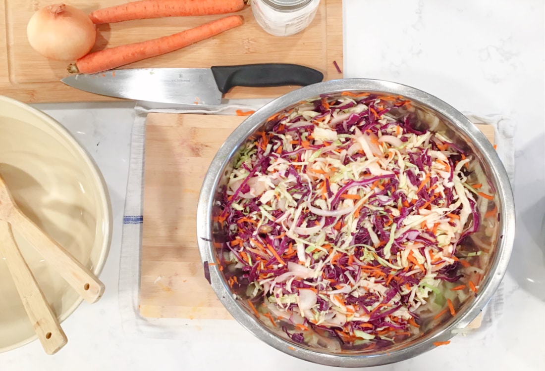 Simple Lacto-Fermented Sauerkraut - Life Full and Frugal - large bowl full of chopped cabbage, onions, and carrots surrounded by bowls, a knife, cutting board and vegetables to make sauerkraut