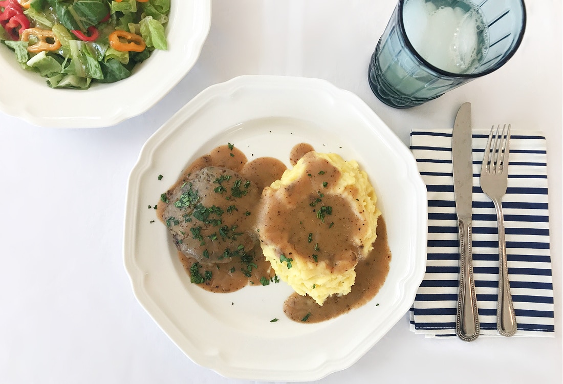 hamburger steaks with mashed potatoes and gravy served with a green salad with peppers set with a white table cloth, blue drinking glass, and a blue and white striped cloth napkin - Frugal Winter Comfort Food: Hamburger Steaks with Mashed Potatoes and Gravy - Life Full and Frugal