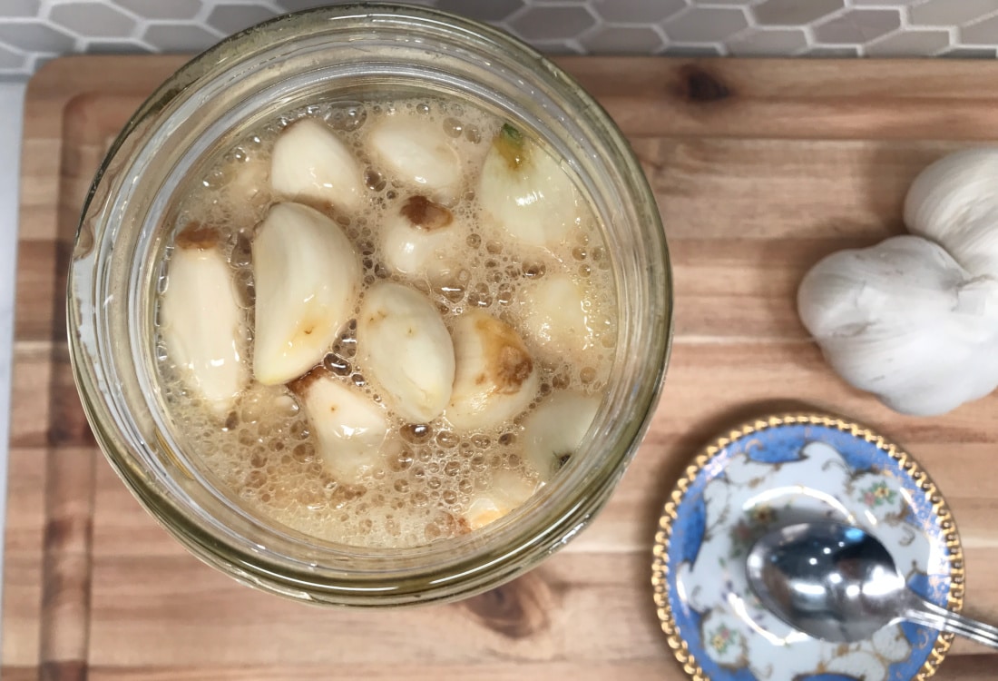 mason jar filled with garlic cloves bubbly from lacto-fermentation in raw honey, placed on a cutting board with heads of garlic and a spoon resting on a vintage piece of china - Lacto-Fermented Garlic Cloves in Raw Honey - Life Full and Frugal