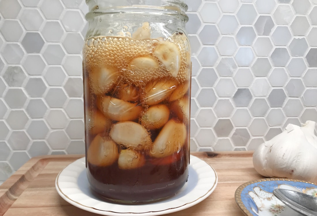 mason jar filled with garlic cloves, fermenting in raw, unfiltered honey, resting on a vintage dish with a marble tile backsplash in the background -Lacto-Fermented Garlic Cloves in Raw Honey - Life Full and Frugal