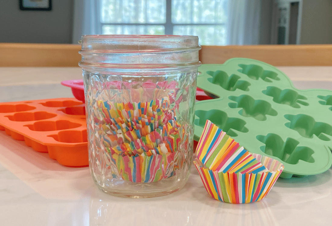 jam jar filled with mini muffin tin paper liners with some on the counter beside it and candy molds in the background - Chocolate Coconut Oil Fudge Candies - Life Full and Frugal
