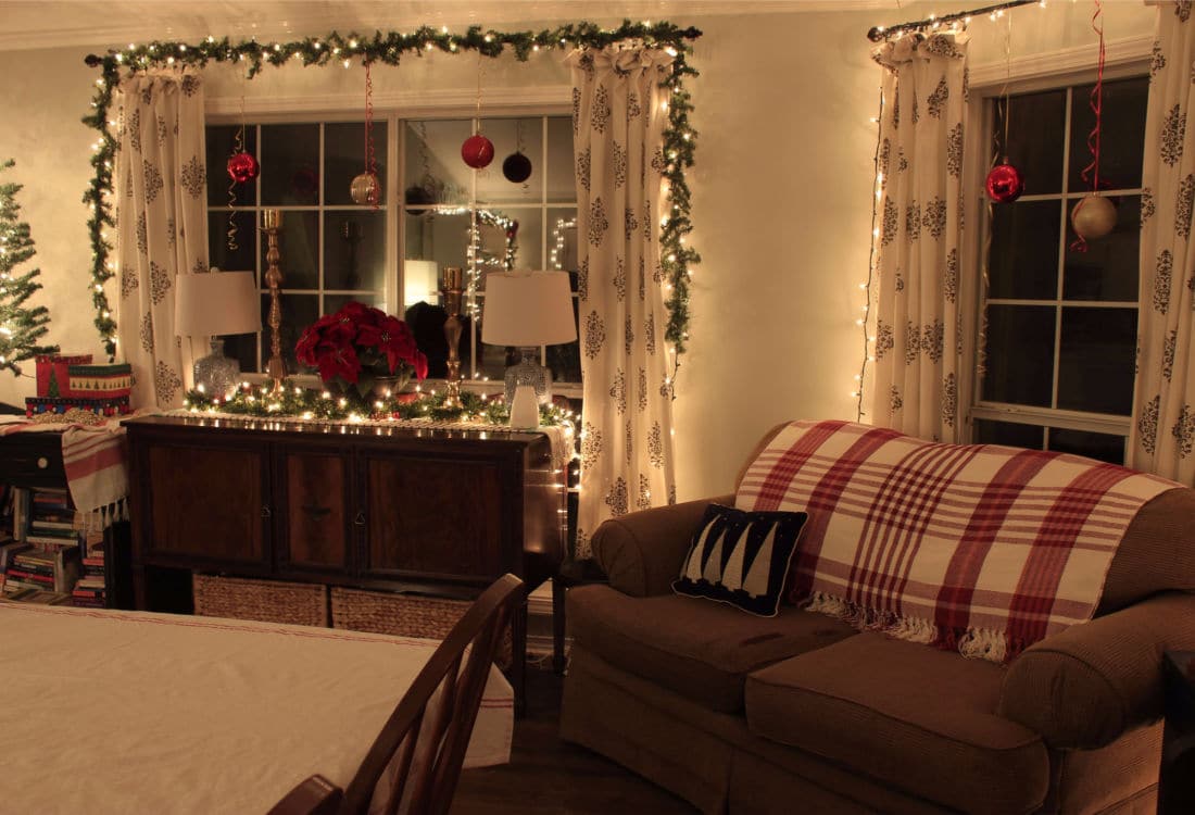 dining room with cream colored table cloth, couch with red and white throw blanket draped over the back, side board covered in Christmas decor in front of a window framed with Christmas lights and garland - 10 Tips for Frugal Christmas Decor