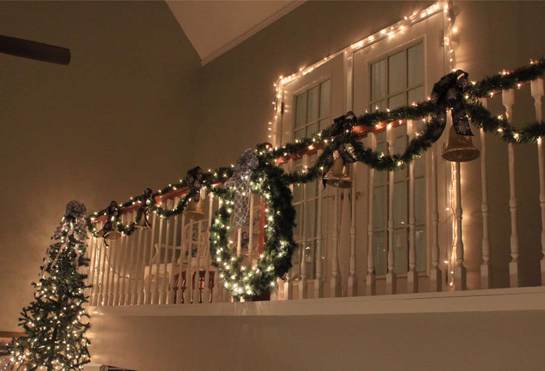 stair banister lit up with Christmas lights and garland with a large wreath hung at the center, doorway in the background framed with Christmas lights and a Christmas tree in the side view - 10 Tips for Frugal Christmas Decor - Life Full and Frugal